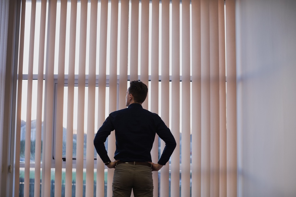 Rear view of man standing with hands on hip near window blinds